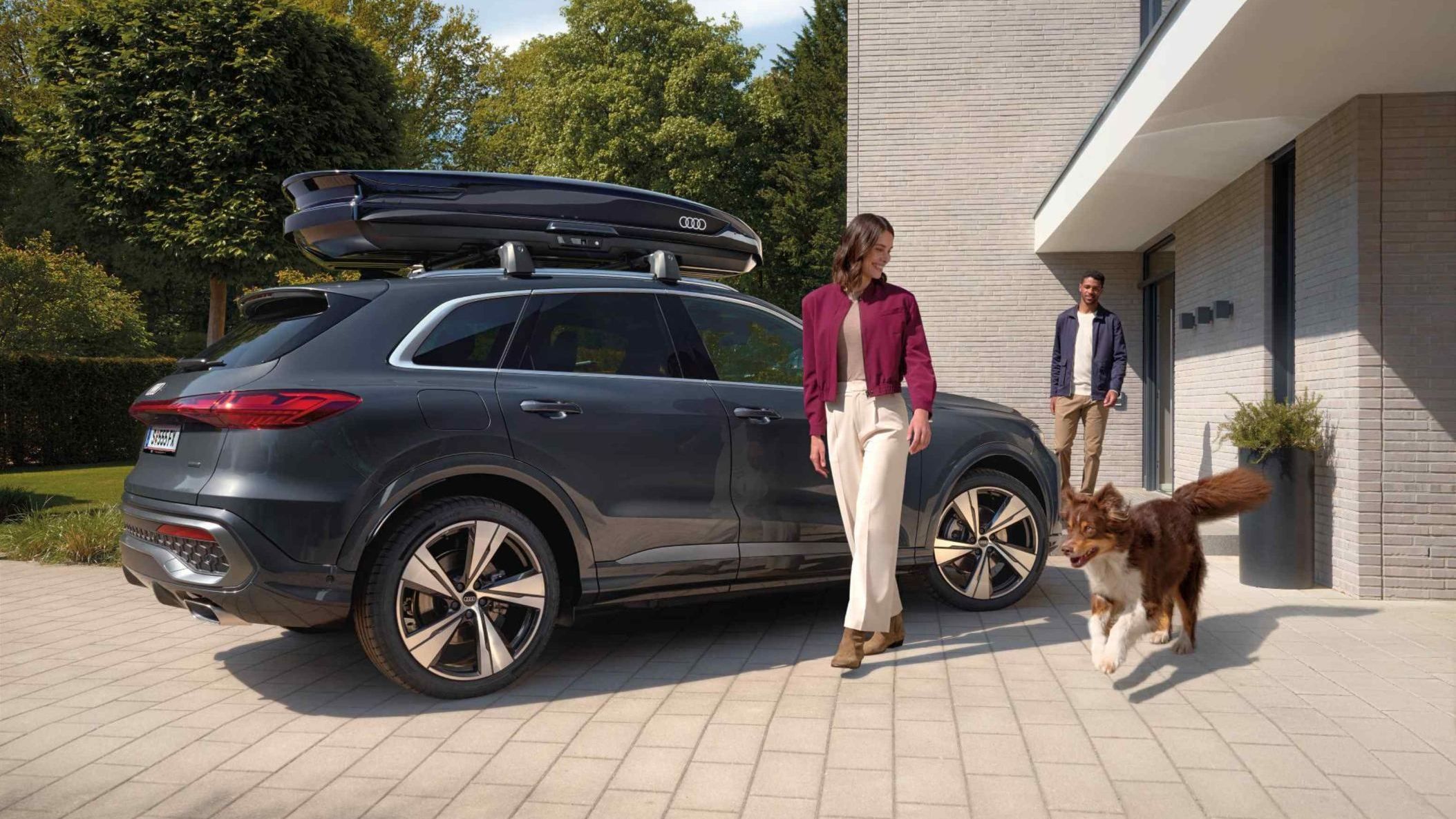 Ein Australian Shepard läuft freudig neben einer lächelnden Frau und einem parkenden Audi Q5 mit Dachbox umher während ein Mann vor dem Hauseingang hinter den beiden steht