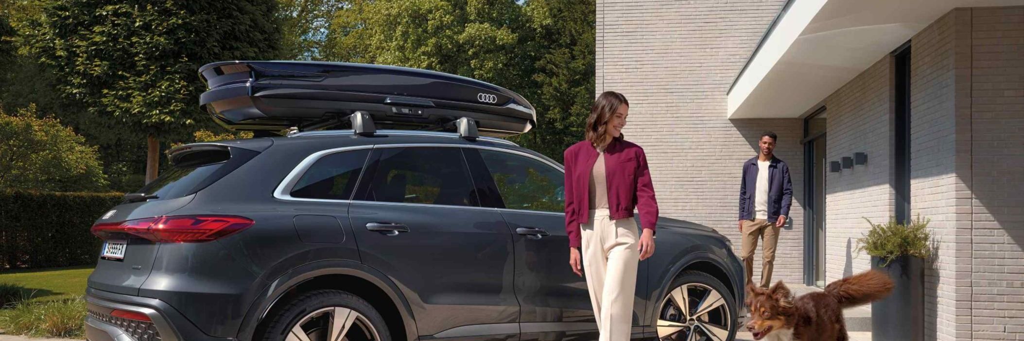 Ein Australian Shepard läuft freudig neben einer lächelnden Frau und einem parkenden Audi Q5 mit Dachbox umher während ein Mann vor dem Hauseingang hinter den beiden steht