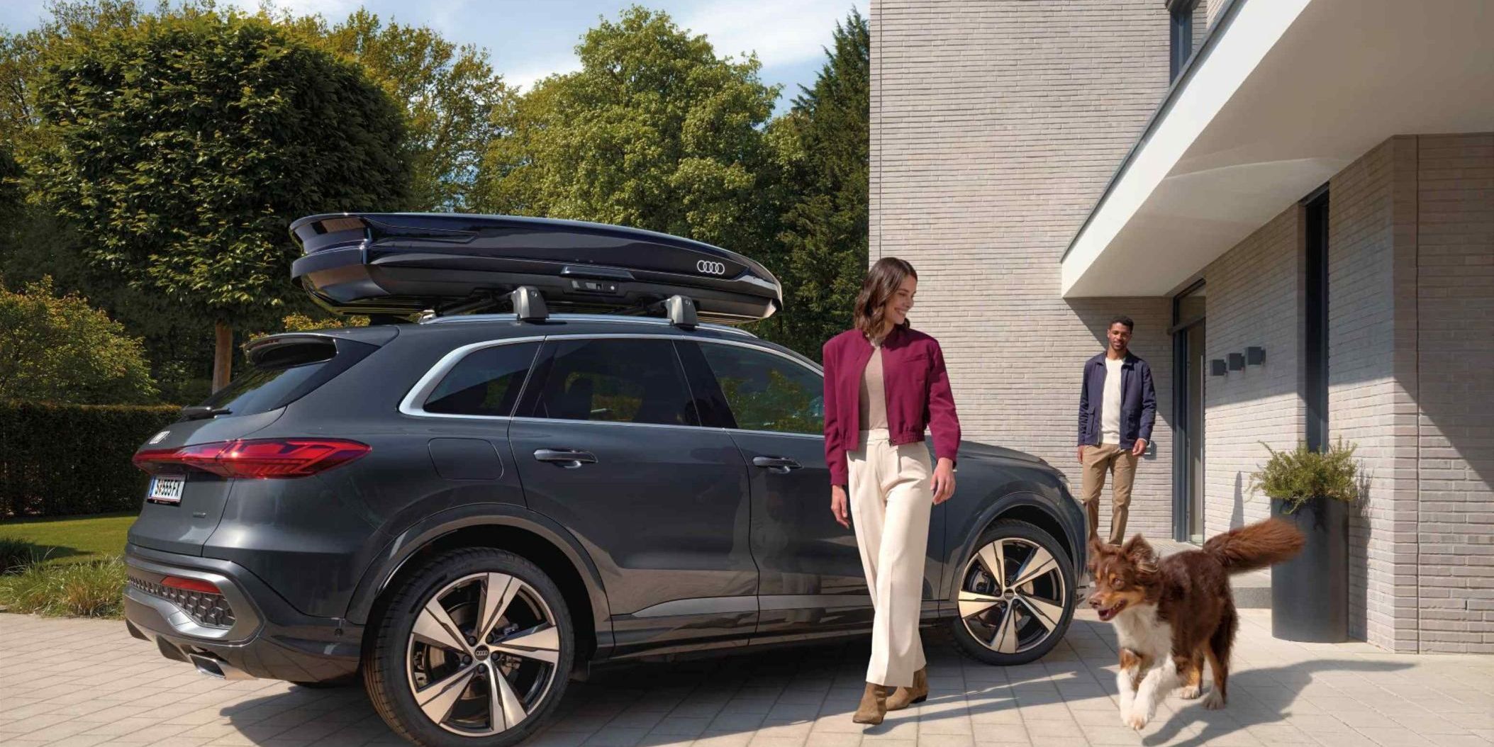 Ein Australian Shepard läuft freudig neben einer lächelnden Frau und einem parkenden Audi Q5 mit Dachbox umher während ein Mann vor dem Hauseingang hinter den beiden steht